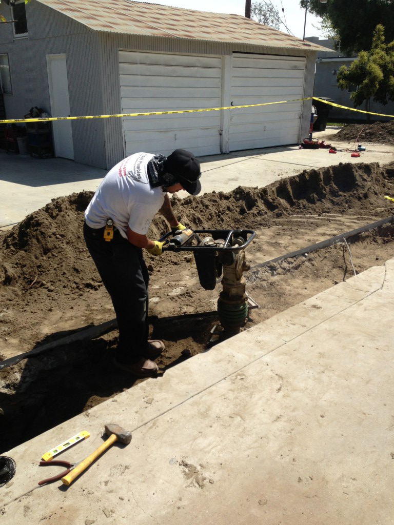 Worker in white shirt installing underground pipe in excavated trench next to garage