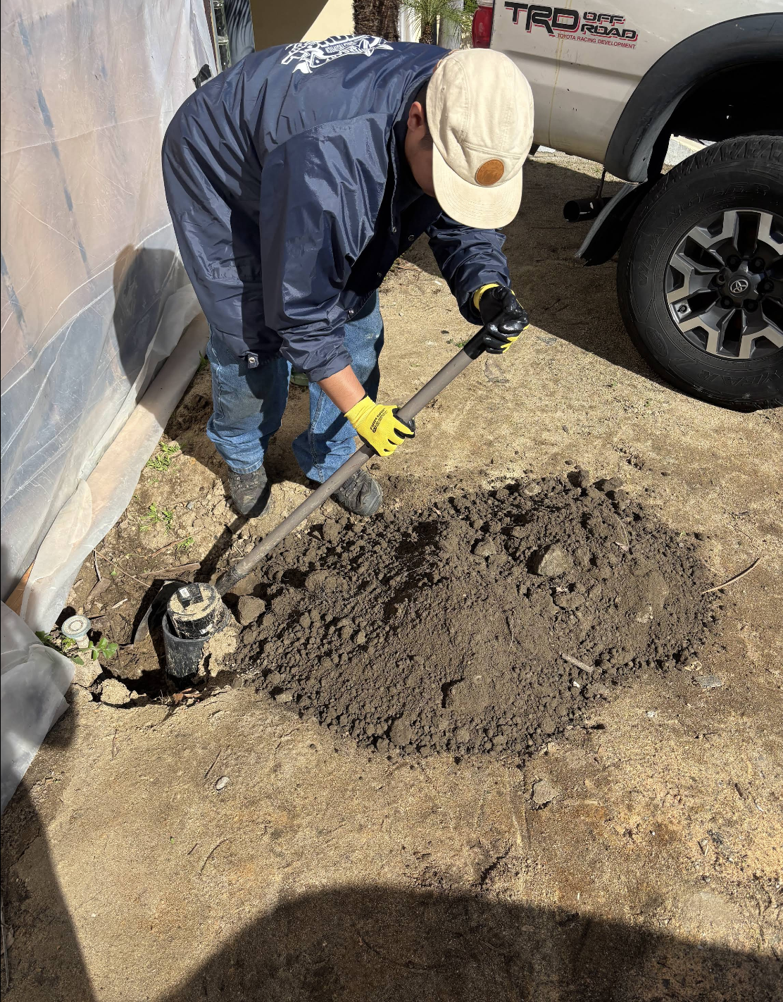 Worker in blue jacket and cream cap using shovel to excavate soil near white trailer and truck wheel